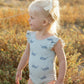 A young child with blond hair in a ponytail wears the Rylee & Cru Baby Girls' & Girls' Scoop Back Onepiece, a light blue swimsuit with whale prints and ruffled sleeves, standing outdoors in natural sunlight.