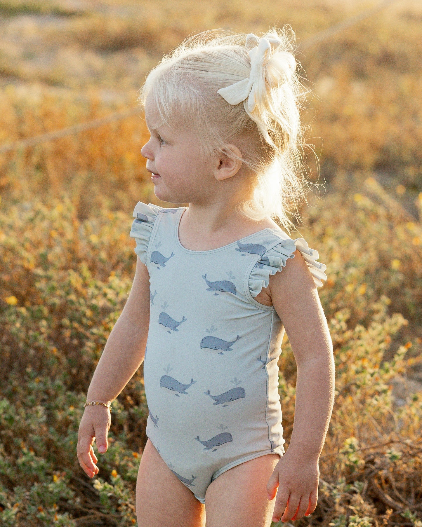 A young child with blond hair in a ponytail wears the Rylee & Cru Baby Girls' & Girls' Scoop Back Onepiece, a light blue swimsuit with whale prints and ruffled sleeves, standing outdoors in natural sunlight.
