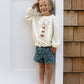 Barefoot child smiling and saluting in a cream sweatshirt with lighthouse graphic, wearing Rylee & Cru Baby Boys' & Boys' Swim Trunk—blue with anchor patterns—standing by a white wall and wood siding.