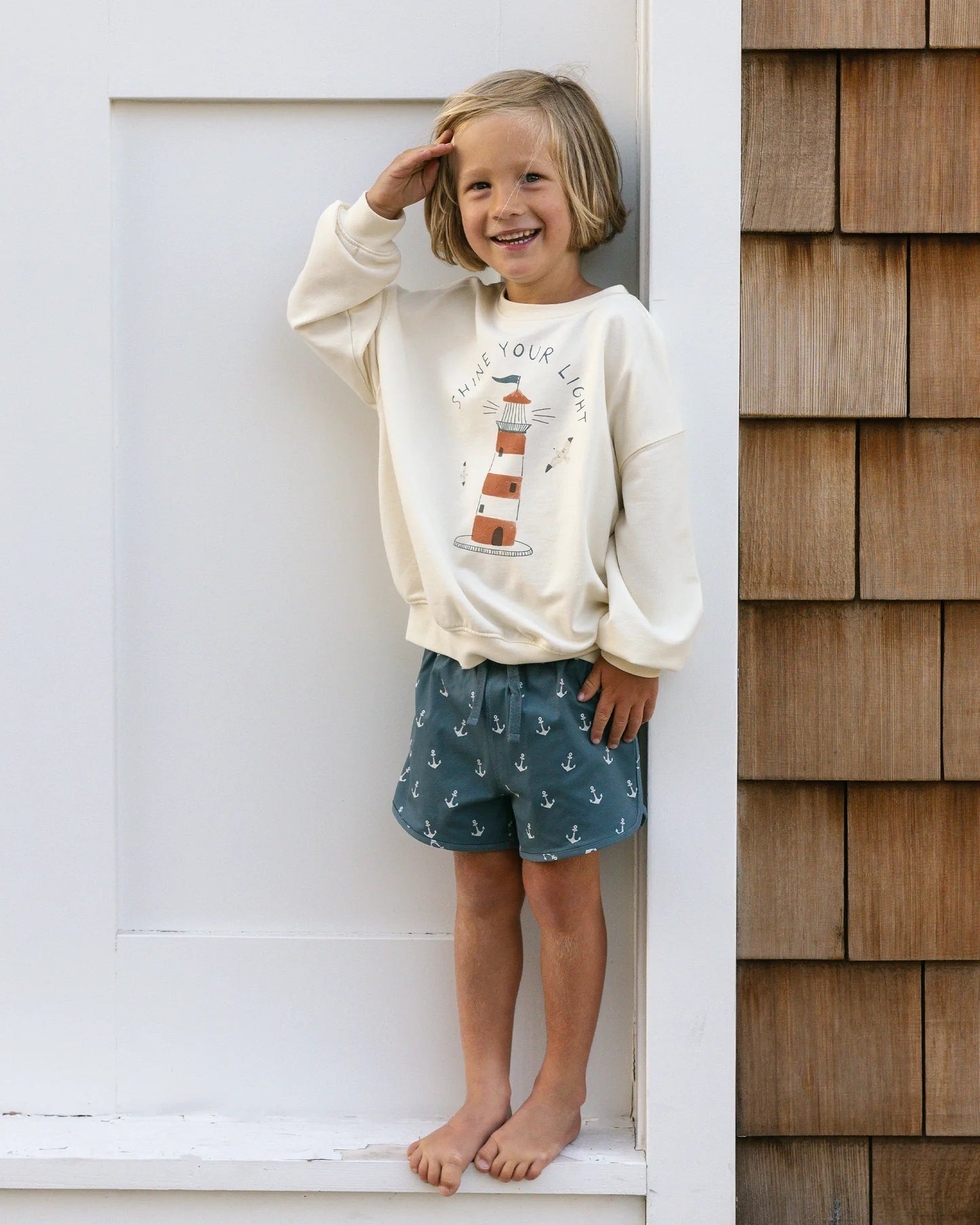 Barefoot child smiling and saluting in a cream sweatshirt with lighthouse graphic, wearing Rylee & Cru Baby Boys' & Boys' Swim Trunk—blue with anchor patterns—standing by a white wall and wood siding.