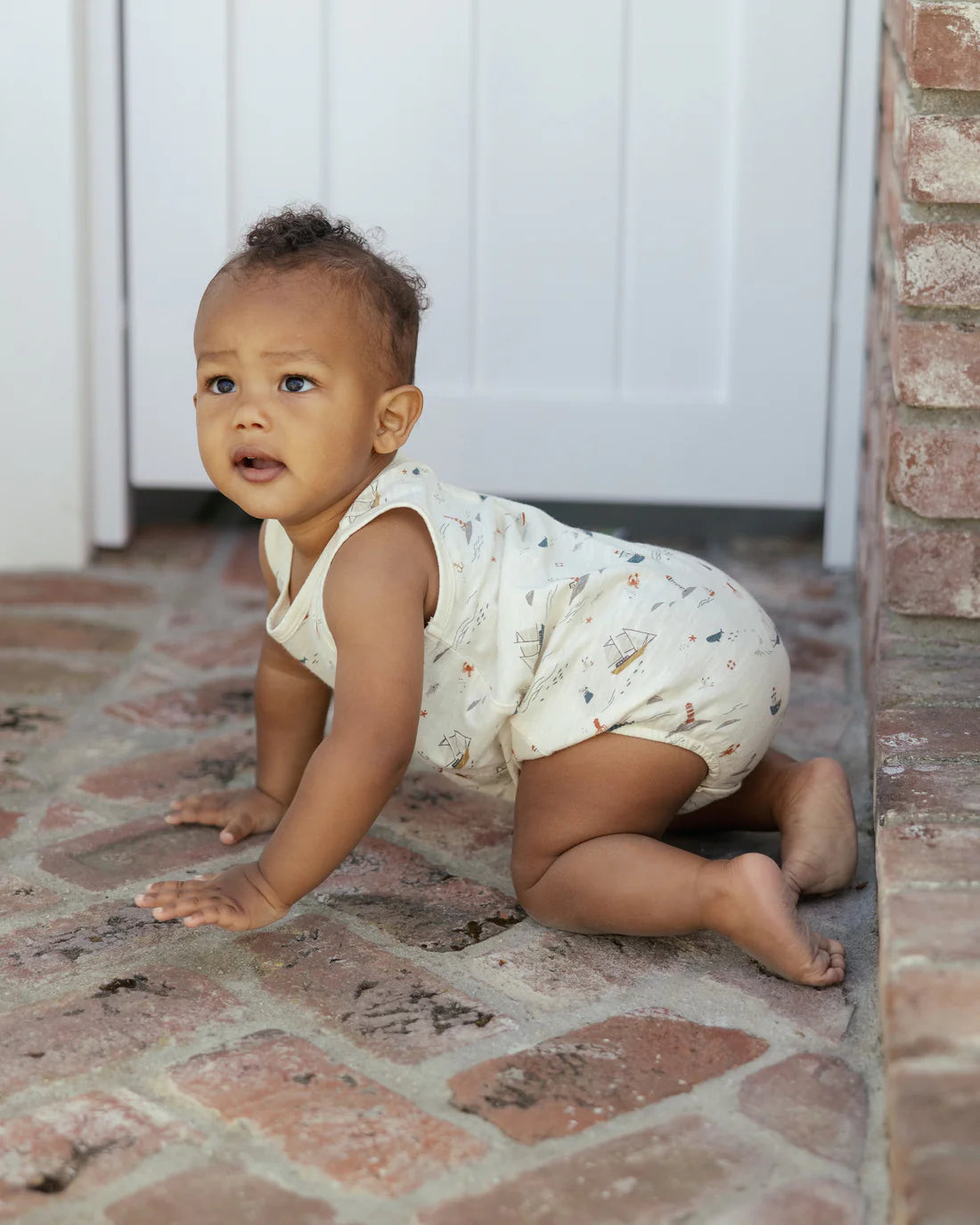 A baby wearing the Rylee & Cru Baby Boys' Bubble Onesie with nautical print crawls on a brick floor near a white door and brick wall.