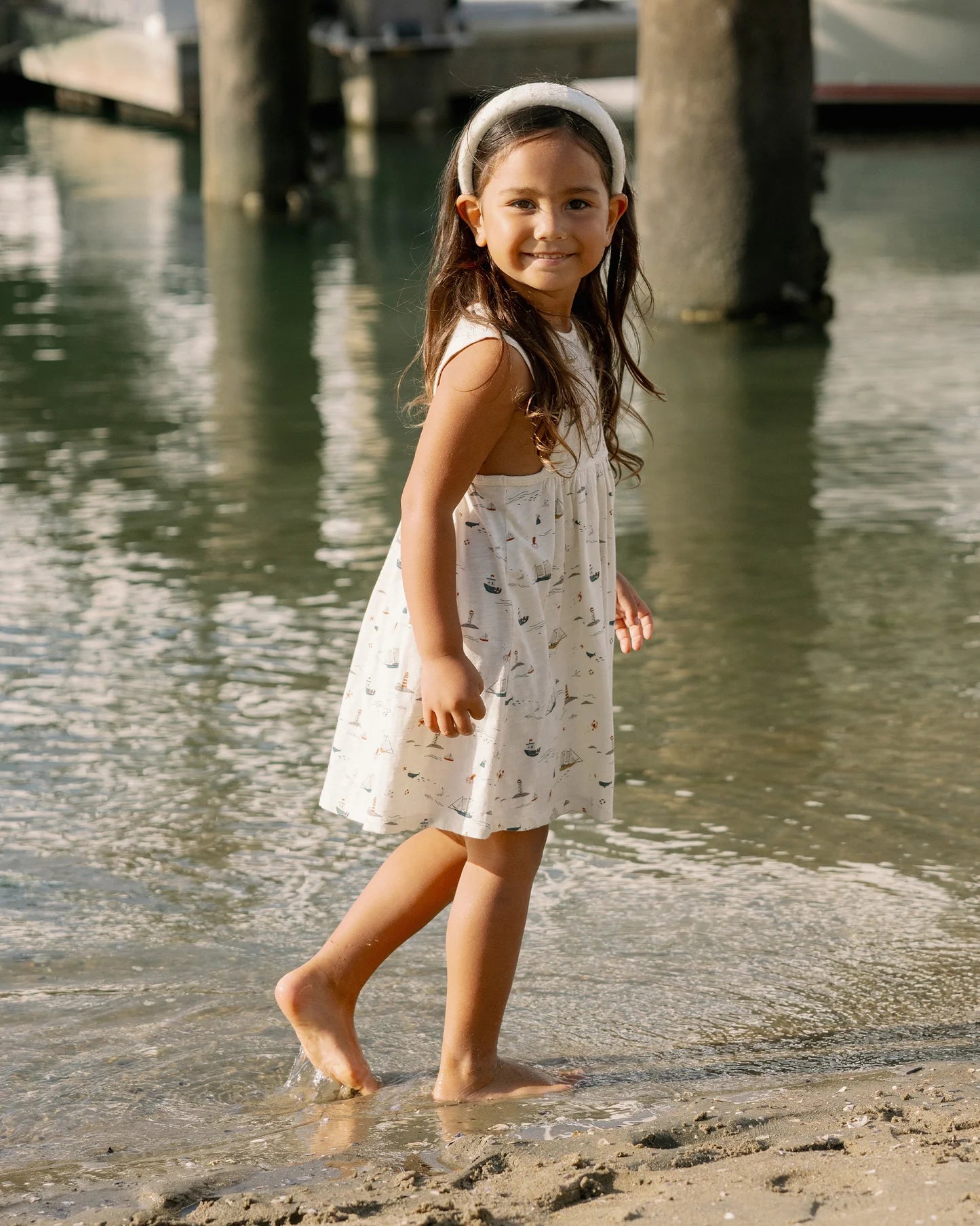 A young girl in the Rylee & Cru Baby Girls' & Girls' Layla Dress smiles barefoot by calm water near a dock.