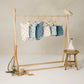 A wooden rack showcases six pastel baby outfits with nautical patterns, including the Rylee & Cru Baby Girls' & Girls' Scoop Back Onepiece swimsuit with ruffled sleeves and UPF 50+. Nearby are a fishing net, rope, toy sailboat, stool, and straw hat.