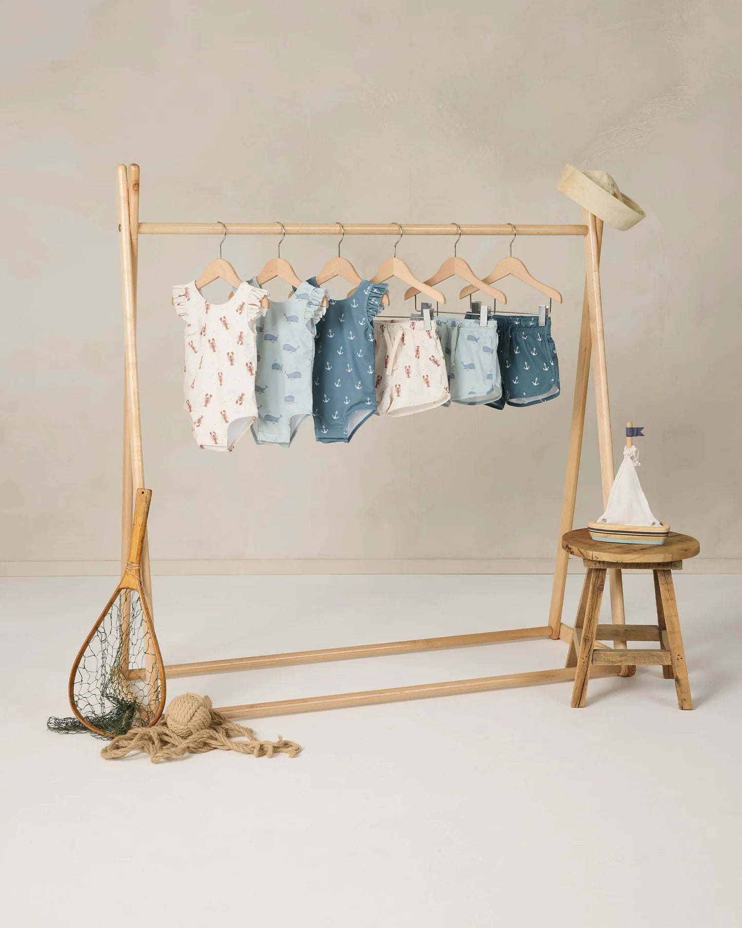 A wooden rack showcases six pastel baby outfits with nautical patterns, including the Rylee & Cru Baby Girls' & Girls' Scoop Back Onepiece swimsuit with ruffled sleeves and UPF 50+. Nearby are a fishing net, rope, toy sailboat, stool, and straw hat.