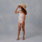 A young girl stands barefoot on a gray surface, smiling to the side while wearing the Rylee & Cru Girls' Scrunch One-Piece swimsuit and a straw hat.