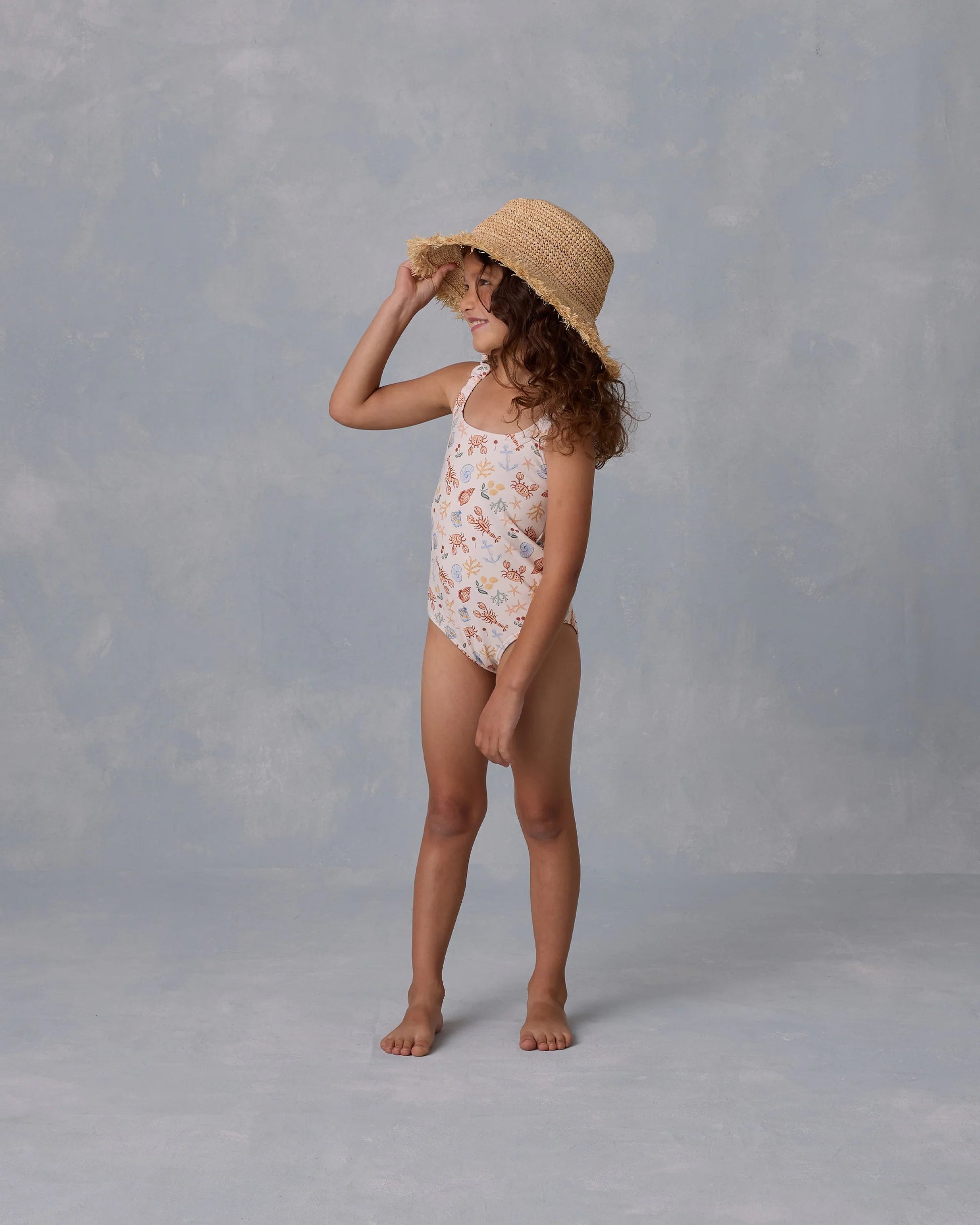A young girl stands barefoot on a gray surface, smiling to the side while wearing the Rylee & Cru Girls' Scrunch One-Piece swimsuit and a straw hat.