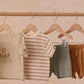 Four pieces of children's clothing, including Rylee & Cru's Boys' Sam Short, hang on wooden hangers: a beige graphic tee, striped tee, eucalyptus green drawstring shorts, and brown shorts—all displayed on a wooden rod.