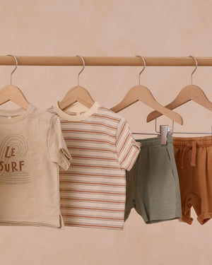 Four pieces of children's clothing, including Rylee & Cru's Boys' Sam Short, hang on wooden hangers: a beige graphic tee, striped tee, eucalyptus green drawstring shorts, and brown shorts—all displayed on a wooden rod.