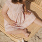 A young child in a pink Rylee & Cru Baby Girls' and Girls' Layla Dress sits on a woven mat at the beach, with a white starfish-shaped toy nearby.