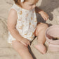 A toddler in the Rylee & Cru Baby Girls' Bubble Onesie, made from soft cotton jersey with a mermaids print, sits on sand near a pink bucket and star-shaped sand toy, looking down.