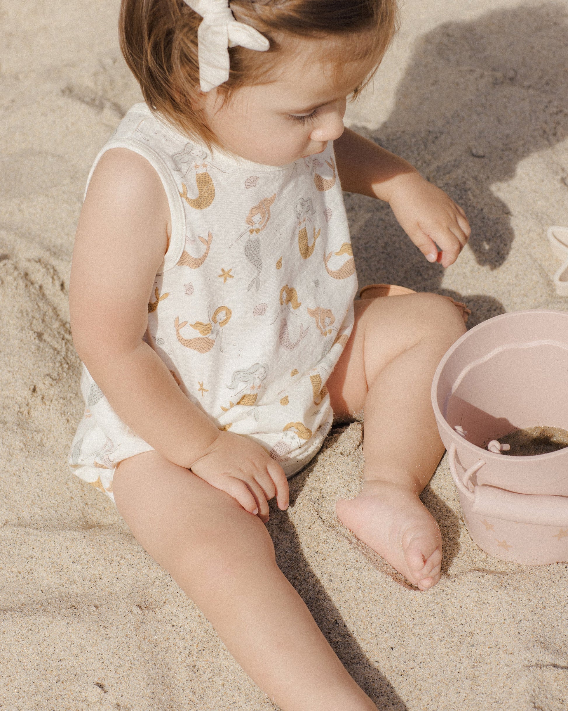A toddler in the Rylee & Cru Baby Girls' Bubble Onesie, made from soft cotton jersey with a mermaids print, sits on sand near a pink bucket and star-shaped sand toy, looking down.