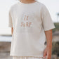A boy wears the Rylee & Cru Baby Boys' and Boys' Raw Edge Tee in light beige cotton slub, featuring "Le Surf" and a wave graphic, while standing outdoors by a rocky beach.