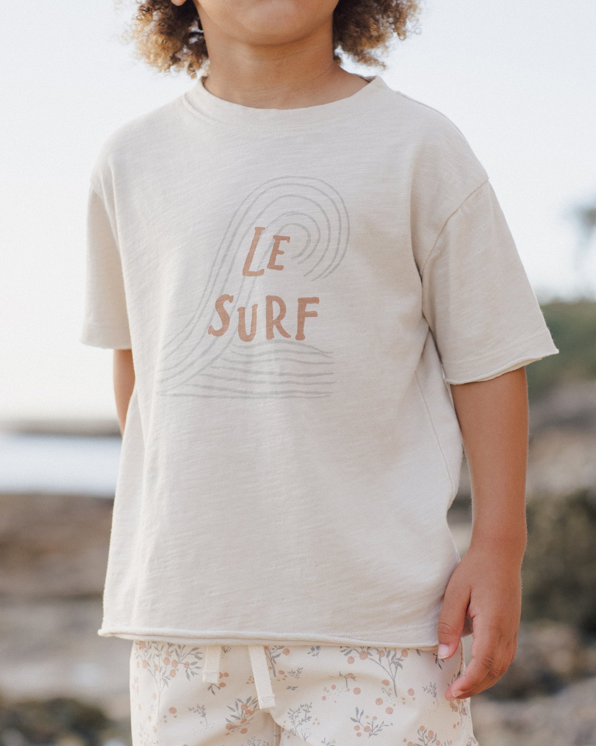 A boy wears the Rylee & Cru Baby Boys' and Boys' Raw Edge Tee in light beige cotton slub, featuring "Le Surf" and a wave graphic, while standing outdoors by a rocky beach.