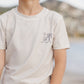 A child wears the Rylee & Cru Boys' Rashguard in light beige with a palm tree and sun graphic, standing outdoors near hills and water—ideal for beach or pool play with UPF 50+ protection.
