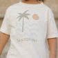 A child wears the Rylee & Cru Baby Boys' and Boys' Raw Edge Tee, made of beige cotton slub fabric, featuring a palm tree, sun, waves graphic, and "Santorini" printed on the front.