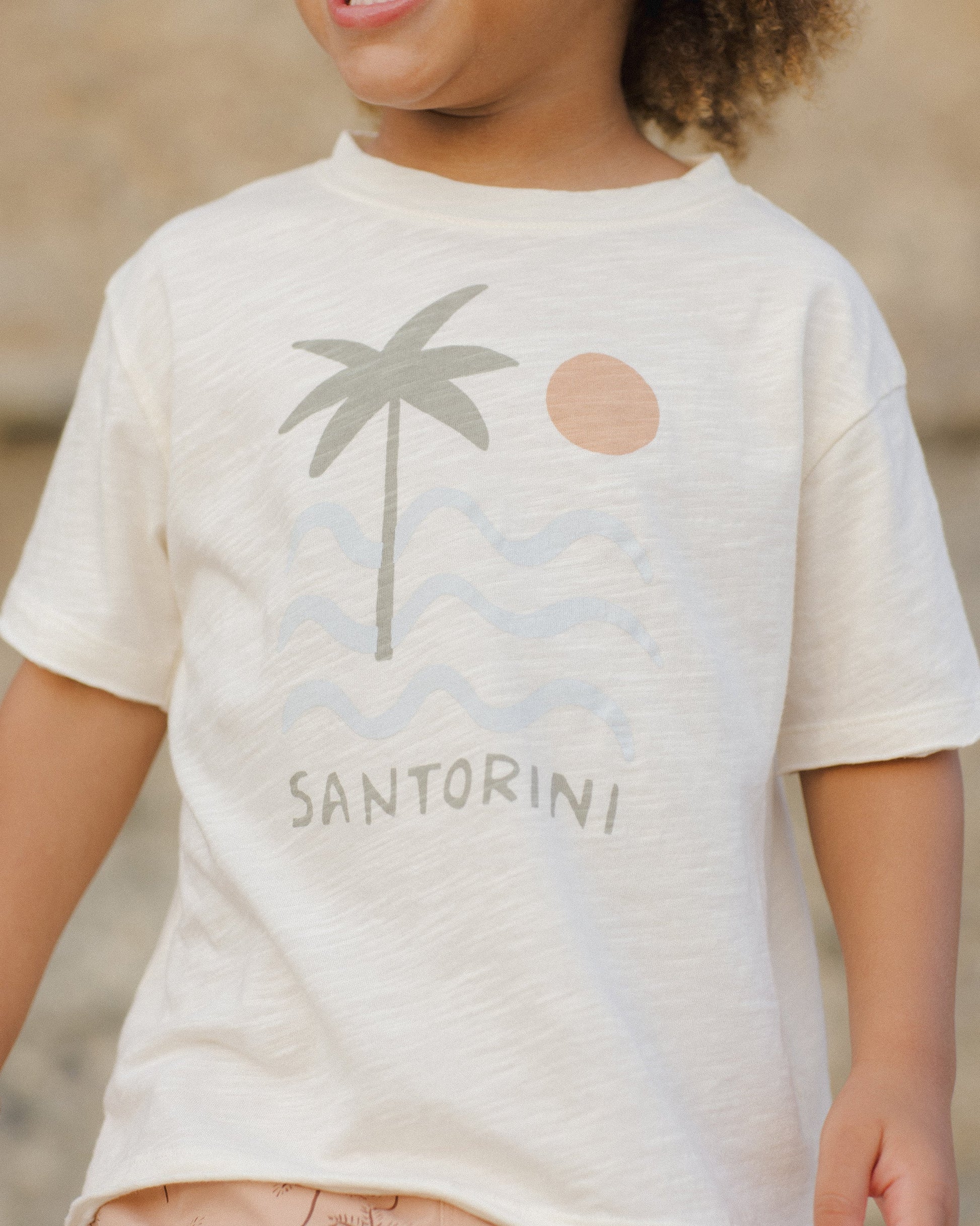 A child wears the Rylee & Cru Baby Boys' and Boys' Raw Edge Tee, made of beige cotton slub fabric, featuring a palm tree, sun, waves graphic, and "Santorini" printed on the front.