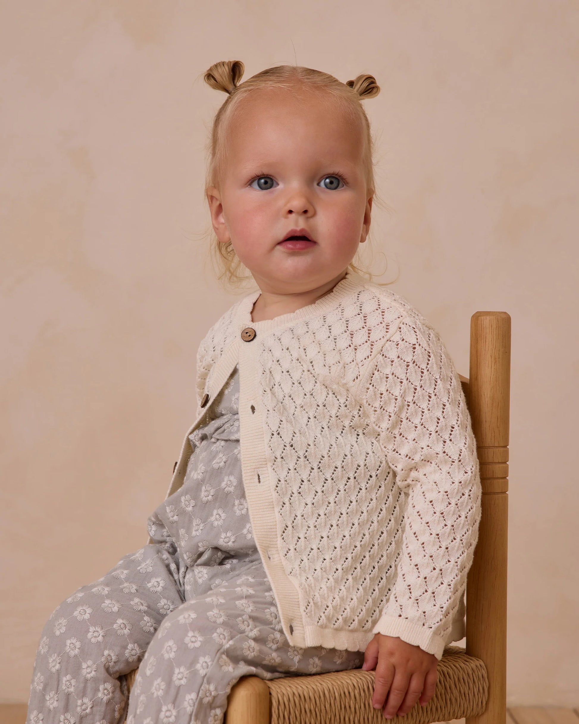 A young blonde girl with pigtails sits on a wooden chair, wearing the Rylee & Cru Baby Girls' Crochet Cardigan and matching pants with a delicate light pattern, looking slightly off camera.