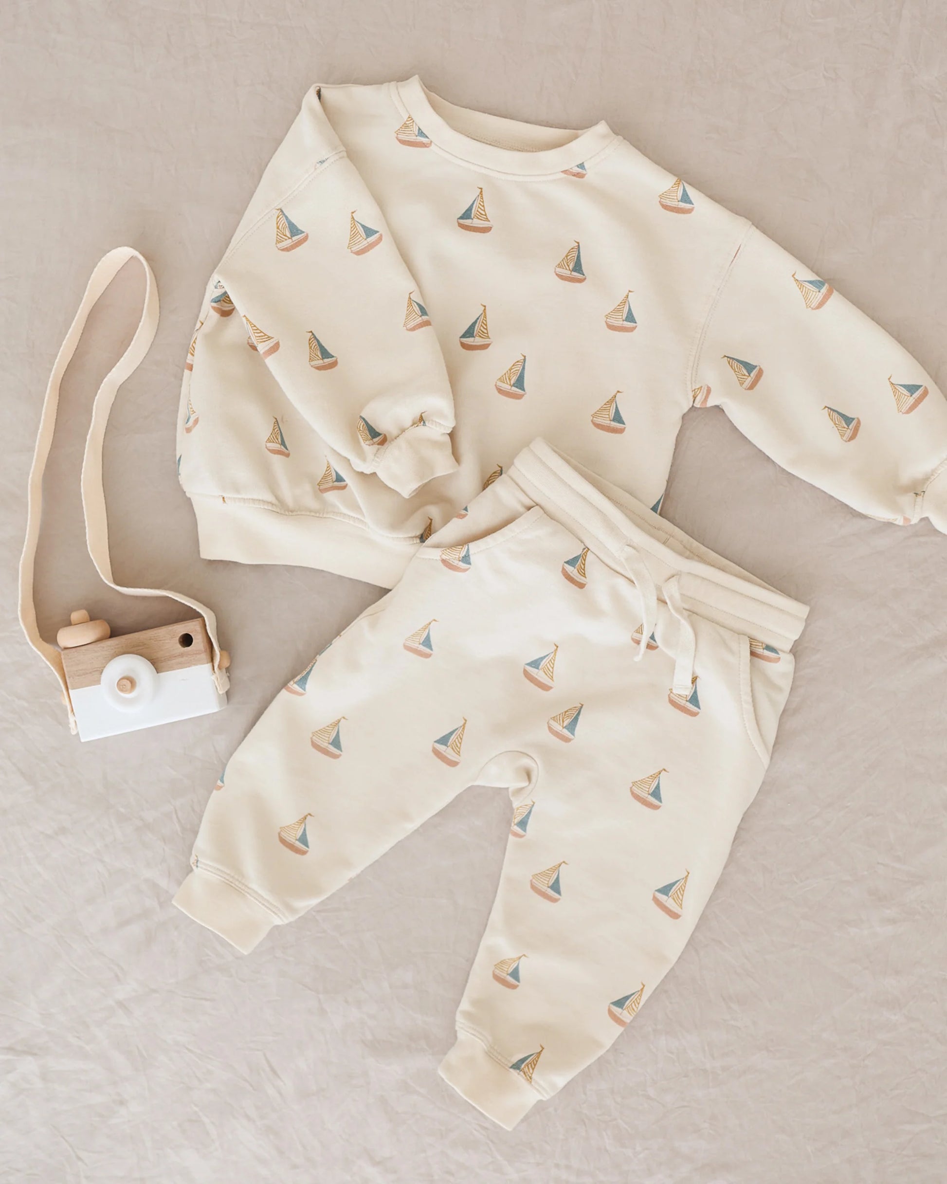 A cream Rylee & Cru Baby Boys' and Boys' Sweatshirt with sailboat print is displayed beside a wooden toy camera with beige strap on a light beige fabric background.