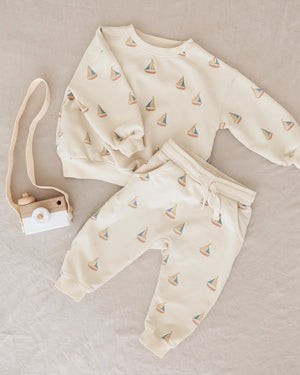 A cream Rylee & Cru Baby Boys' and Boys' Sweatshirt with sailboat print is displayed beside a wooden toy camera with beige strap on a light beige fabric background.