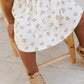 A child in the Rylee & Cru Baby Girls' and Girls' Layla Dress sits on a light wooden chair, visible from the waist down, wearing tan sandals. The dress features a white cotton jersey fabric with a yellow floral pattern.