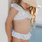 A young child in a Rylee & Cru Girls' Skirted Bikini with an asymmetrical ruffle top and straw hat stands by a white wall near a pool, face partly shaded. UPF 50+ fabric offers sun-safe style for all-day play.