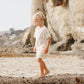 A blond-haired young child wears the Rylee & Cru Baby Boys' and Boys' Play Set while walking barefoot on a stone ledge at the beach, with cliffs and a stone tower in the background, ready for summer adventures.
