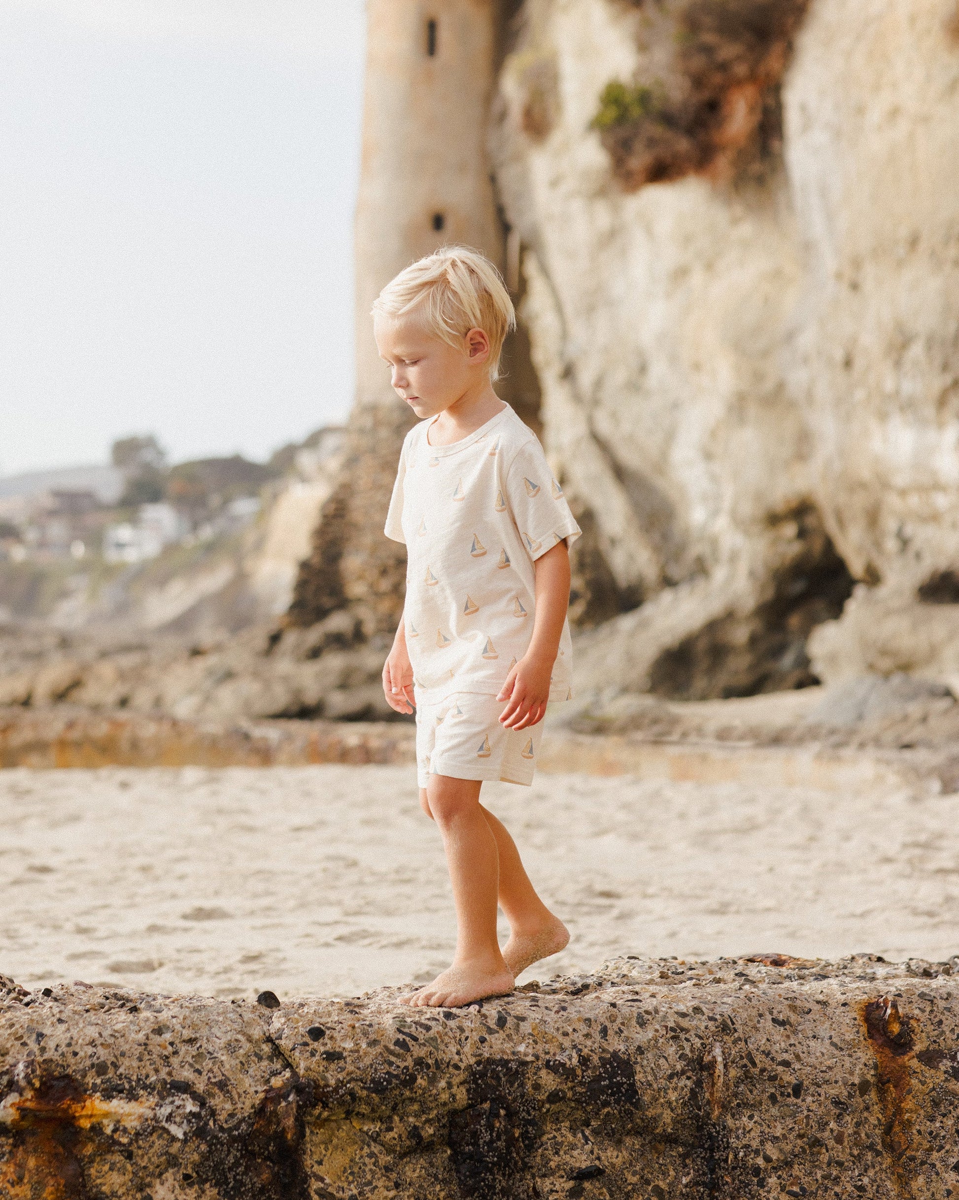 A blond-haired young child wears the Rylee & Cru Baby Boys' and Boys' Play Set while walking barefoot on a stone ledge at the beach, with cliffs and a stone tower in the background, ready for summer adventures.