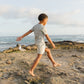 A child wears the Rylee & Cru Baby and Kids' Short-Sleeve Rashguard, walking barefoot on rocky ground by the ocean under a cloudy sky, with a seagull in the background.