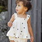A toddler with curly hair stands outdoors near a wooden fence, wearing the Rylee & Cru Baby Girls' Lala Set—a white, ruffled dress with black polka dots and smocked ruffle detail—looking to the side.
