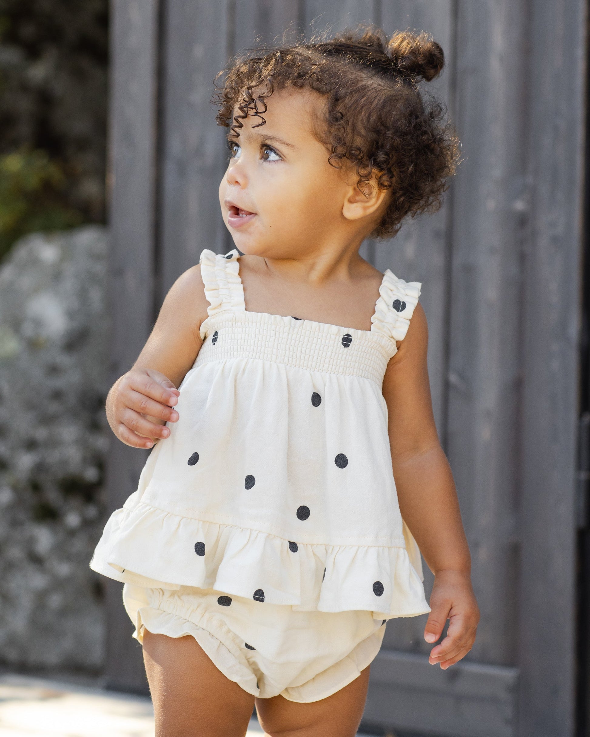 A toddler with curly hair stands outdoors near a wooden fence, wearing the Rylee & Cru Baby Girls' Lala Set—a white, ruffled dress with black polka dots and smocked ruffle detail—looking to the side.