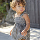 A toddler wears the Rylee & Cru Baby Girls' Julia Romper, featuring black and white gingham with ruffle detail and shoulder straps, standing outdoors with curly hair and a neutral expression.