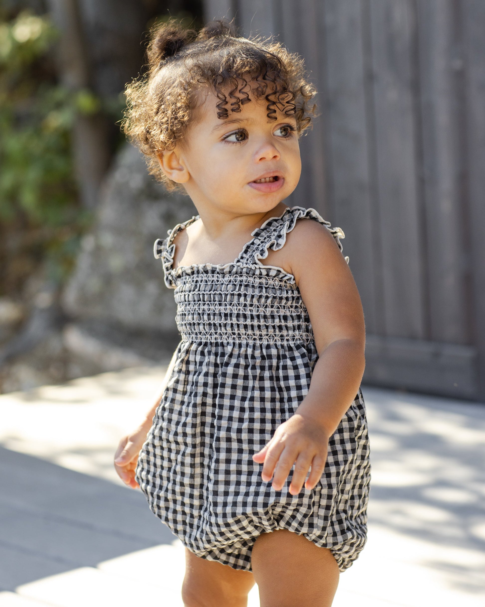A toddler wears the Rylee & Cru Baby Girls' Julia Romper, featuring black and white gingham with ruffle detail and shoulder straps, standing outdoors with curly hair and a neutral expression.