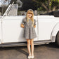A young girl wears the Rylee & Cru Girls' Louise Dress—a black and white checkered style with a gathered skirt and puff sleeves—while standing in front of a classic white convertible on a sunny day.