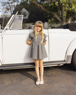 A young girl wears the Rylee & Cru Girls' Louise Dress—a black and white checkered style with a gathered skirt and puff sleeves—while standing in front of a classic white convertible on a sunny day.