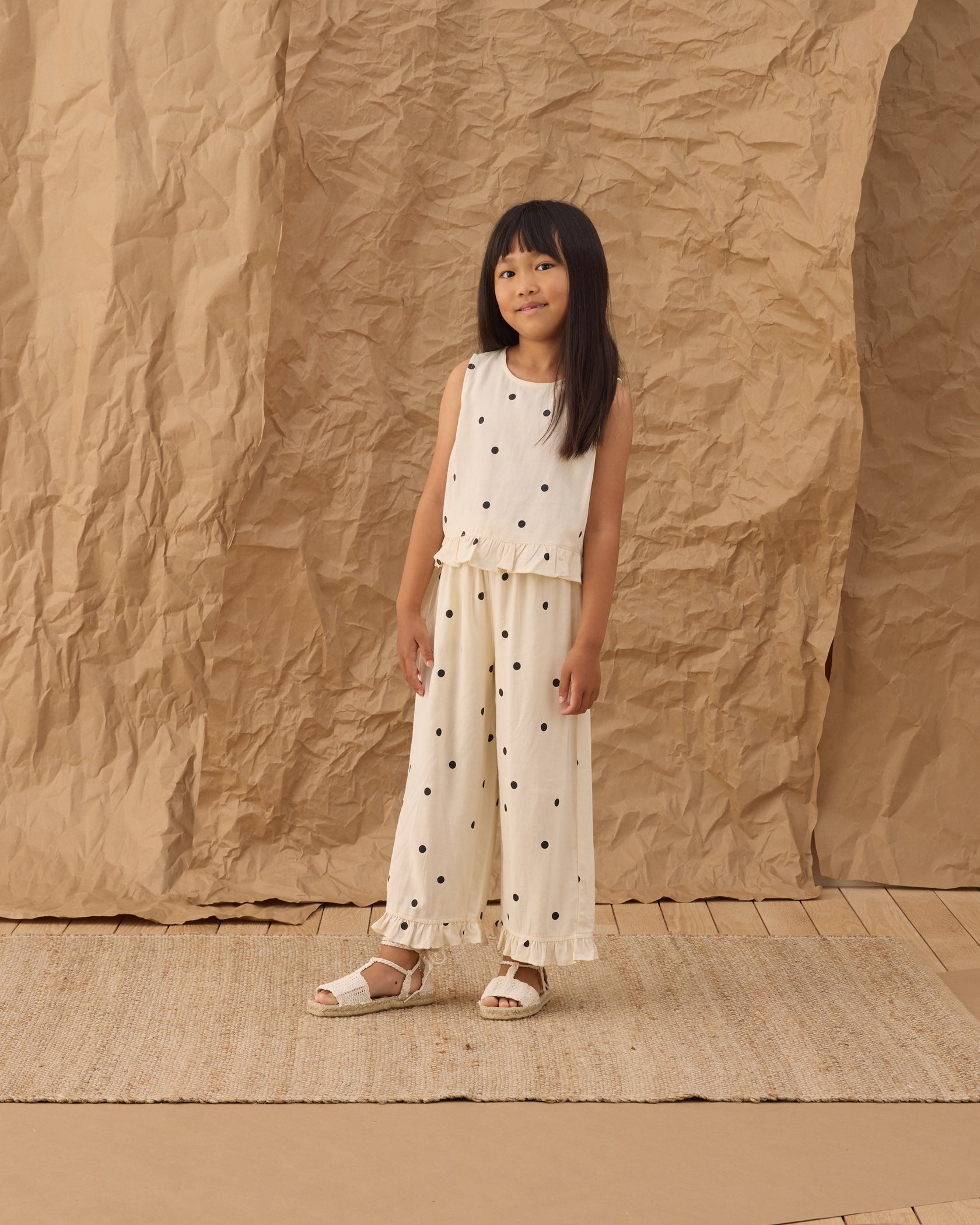 A young girl stands on a woven rug in front of a crumpled brown paper backdrop, wearing the sleeveless cotton Girls' Oceanside Top by Rylee & Cru and wide-leg polka-dot pants, paired with white sandals.