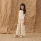 A young girl stands on a woven rug, dressed in Rylee & Cru’s Girls’ Wide Leg Pant—a white, sleeveless, polka-dotted style with an elastic drawstring waist and comfortable silhouette—paired with sandals and set against a crumpled brown paper backdrop.