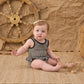 A baby wearing the Rylee & Cru Baby Girls' Skirted One-Piece in a classic black-and-white checkered pattern sits on a woven mat, holding a net, with a ship’s wheel and crumpled brown backdrop behind—perfect for poolside adventures thanks to its UPF 50+ protection.