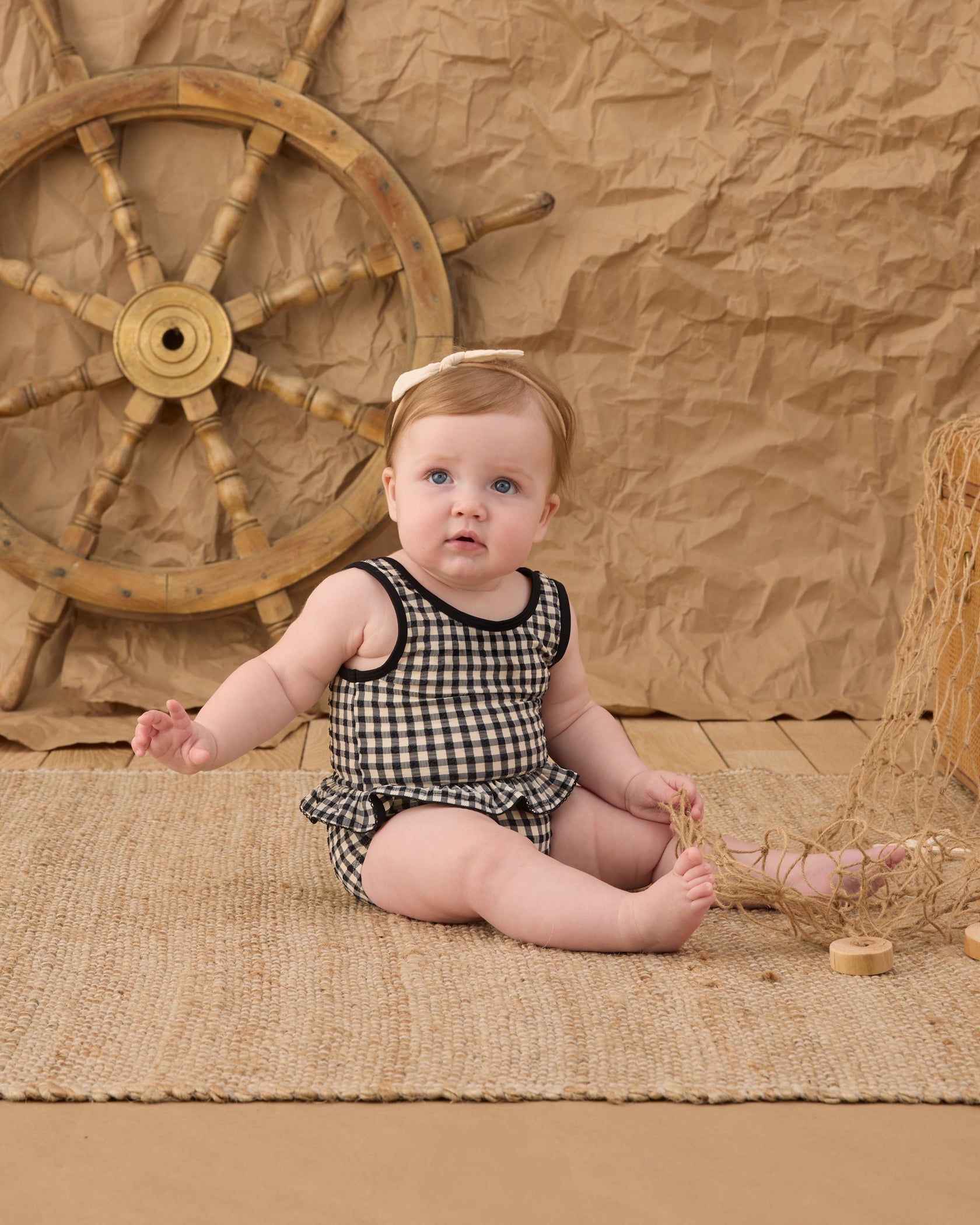 A baby wearing the Rylee & Cru Baby Girls' Skirted One-Piece in a classic black-and-white checkered pattern sits on a woven mat, holding a net, with a ship’s wheel and crumpled brown backdrop behind—perfect for poolside adventures thanks to its UPF 50+ protection.