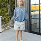 A smiling child stands on a wooden deck in the Rylee & Cru Baby Boys' and Boys' Relaxed Sweatshirt, featuring cozy French terry fabric, "COASTAL" print, blue color, paired with striped shorts, backward cap, and slip-on shoes with hands in pockets.