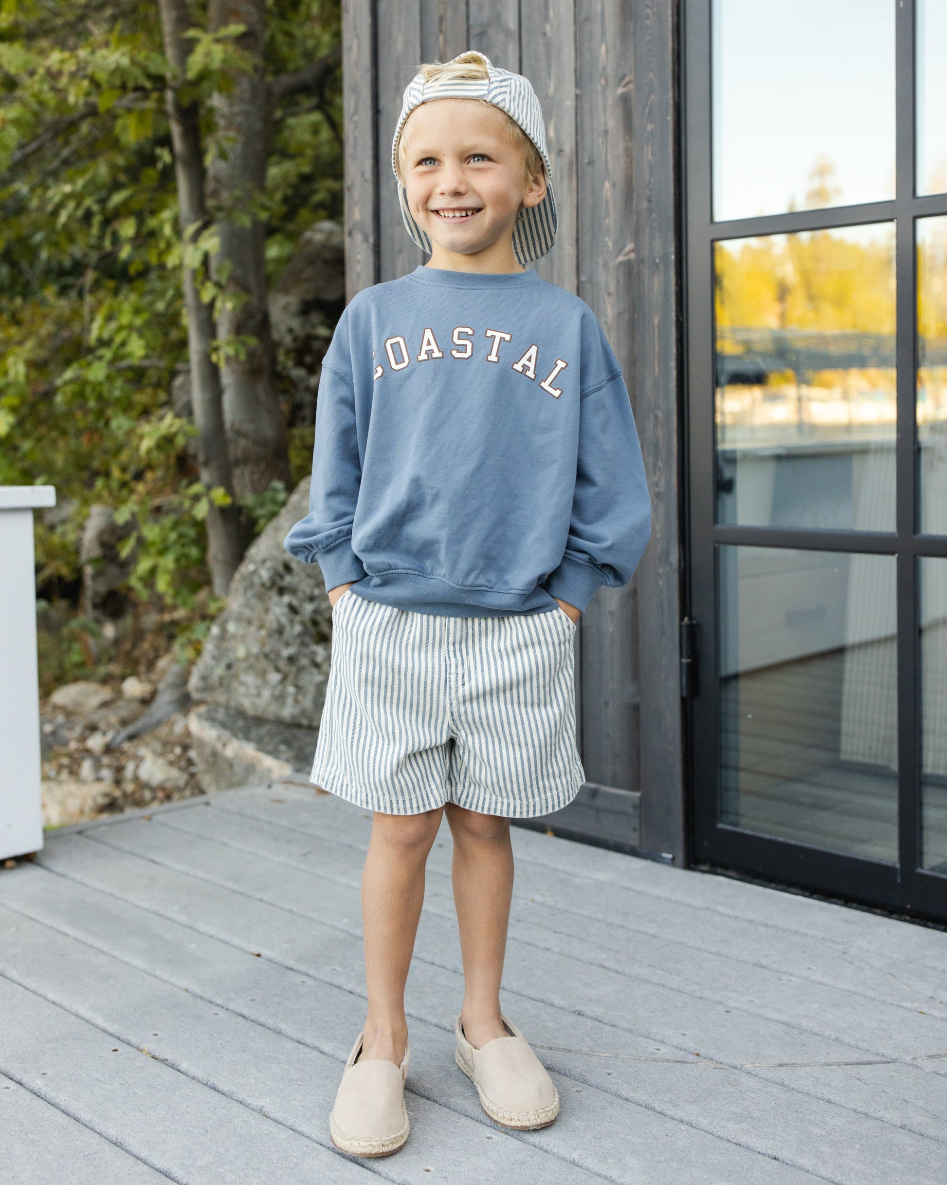 A smiling child stands on a wooden deck in the Rylee & Cru Baby Boys' and Boys' Relaxed Sweatshirt, featuring cozy French terry fabric, "COASTAL" print, blue color, paired with striped shorts, backward cap, and slip-on shoes with hands in pockets.