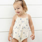 A young child in a Rylee & Cru Baby Girls' Nala Romper—cream cotton jersey with blue shell patterns and bow straps—stands before a white shiplap wall, looking to the side with a smile.
