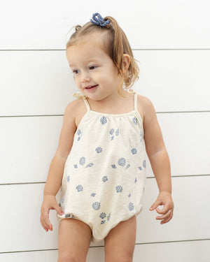 A young child in a Rylee & Cru Baby Girls' Nala Romper—cream cotton jersey with blue shell patterns and bow straps—stands before a white shiplap wall, looking to the side with a smile.