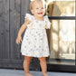 A toddler with blonde hair stands barefoot on a wooden deck in front of a glass door, wearing the Rylee & Cru Baby Girls' and Girls' Mariposa Dress— a cream dress with blue patterns and butterfly sleeves.