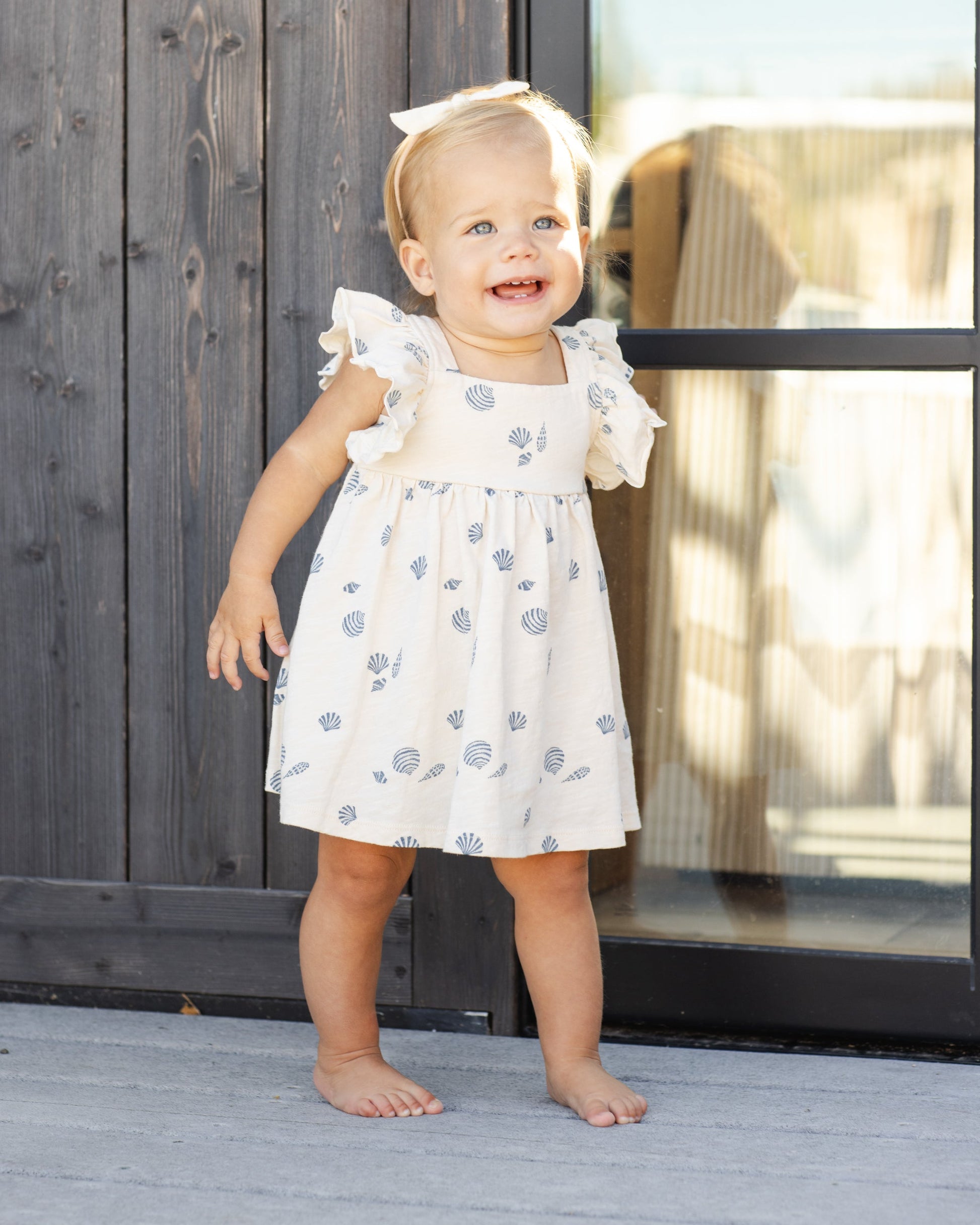 A toddler with blonde hair stands barefoot on a wooden deck in front of a glass door, wearing the Rylee & Cru Baby Girls' and Girls' Mariposa Dress— a cream dress with blue patterns and butterfly sleeves.