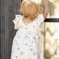 A young child with blond hair wears the Rylee & Cru Baby Girls' and Girls' Mariposa Dress—white with blue shell patterns and butterfly sleeves—while standing by a window, one hand touching the glass.