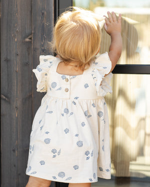 A young child with blond hair wears the Rylee & Cru Baby Girls' and Girls' Mariposa Dress—white with blue shell patterns and butterfly sleeves—while standing by a window, one hand touching the glass.