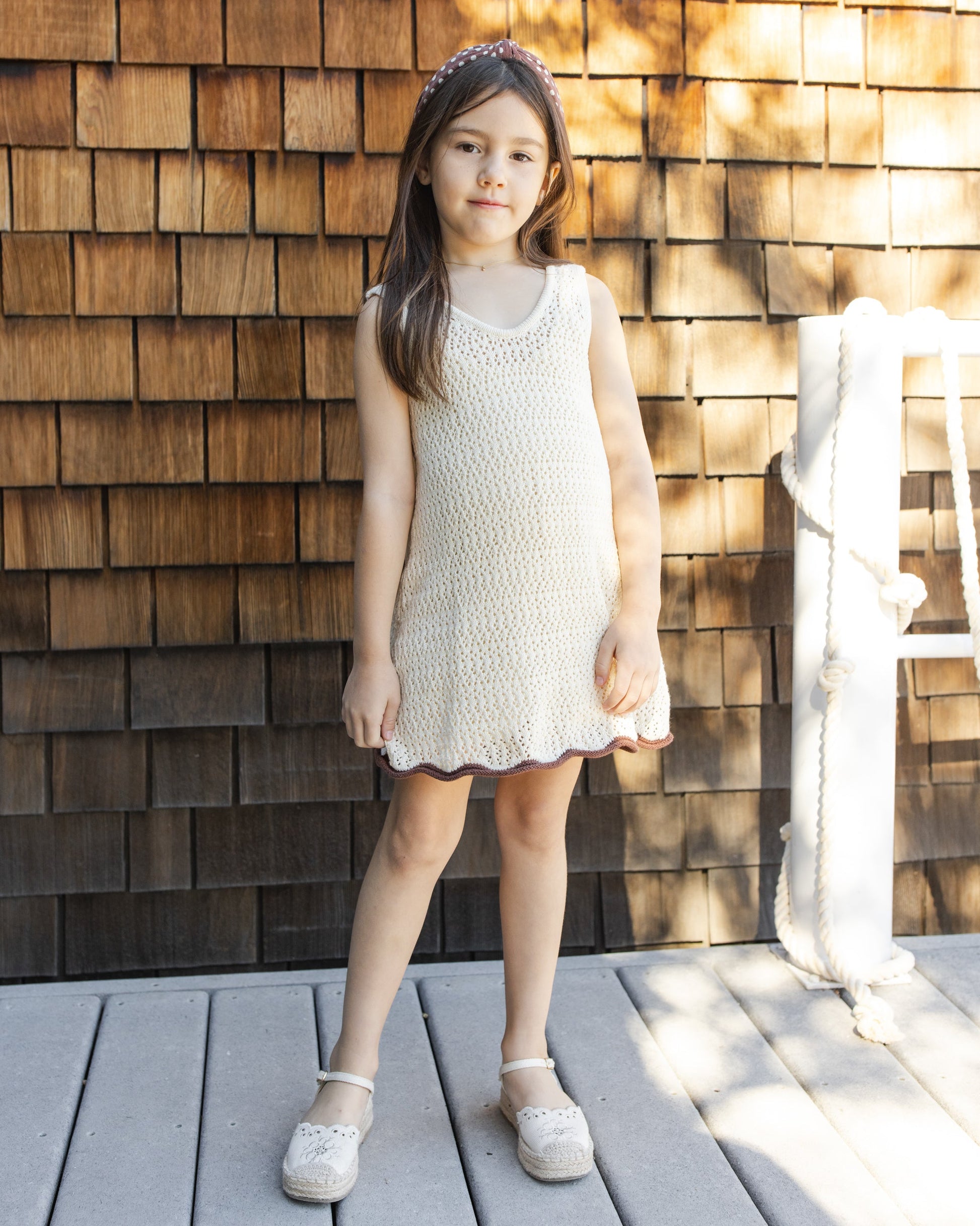 A young girl wears the Rylee & Cru Girls' Crochet Tank Mini Dress, standing on a wooden deck before a shingled wall, with rope and sunlight in the background.