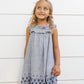 A young girl with curly blonde hair smiles as she stands in front of a white paneled wall, wearing the Girls' Lennon Dress by Rylee & Cru—a blue and white checkered mini dress with a ruffled neckline.