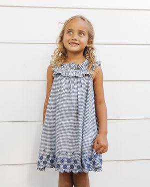 A young girl with curly blonde hair smiles as she stands in front of a white paneled wall, wearing the Girls' Lennon Dress by Rylee & Cru—a blue and white checkered mini dress with a ruffled neckline.
