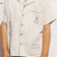A child wears the Rylee & Cru Boys' Lapel Collar Shirt, a short-sleeved cream top with anchor, boat, and fish embroidery, paired with checkered shorts, standing against a white background.